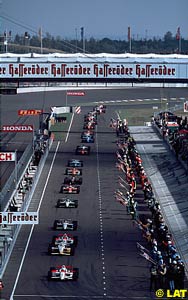 The field is led down pit lane, past the row of flags